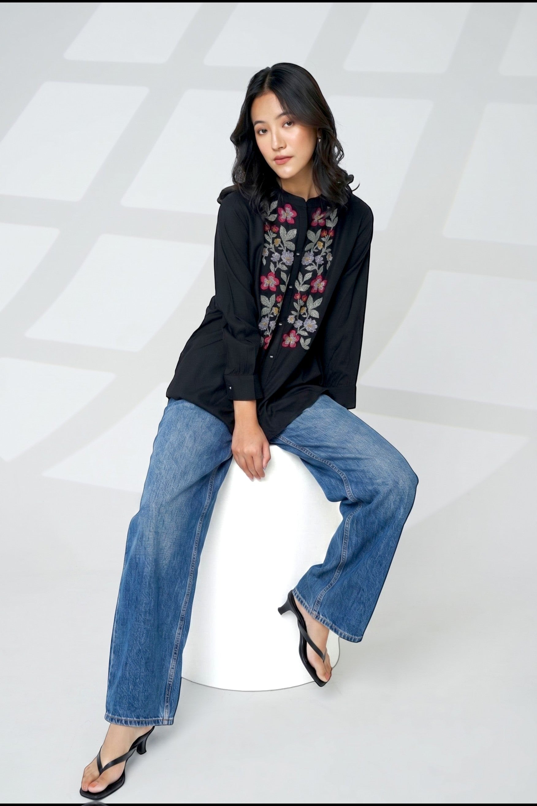 A woman wearing an indigrey black shirt with floral cross-stitch embroidery, paired with blue jeans.