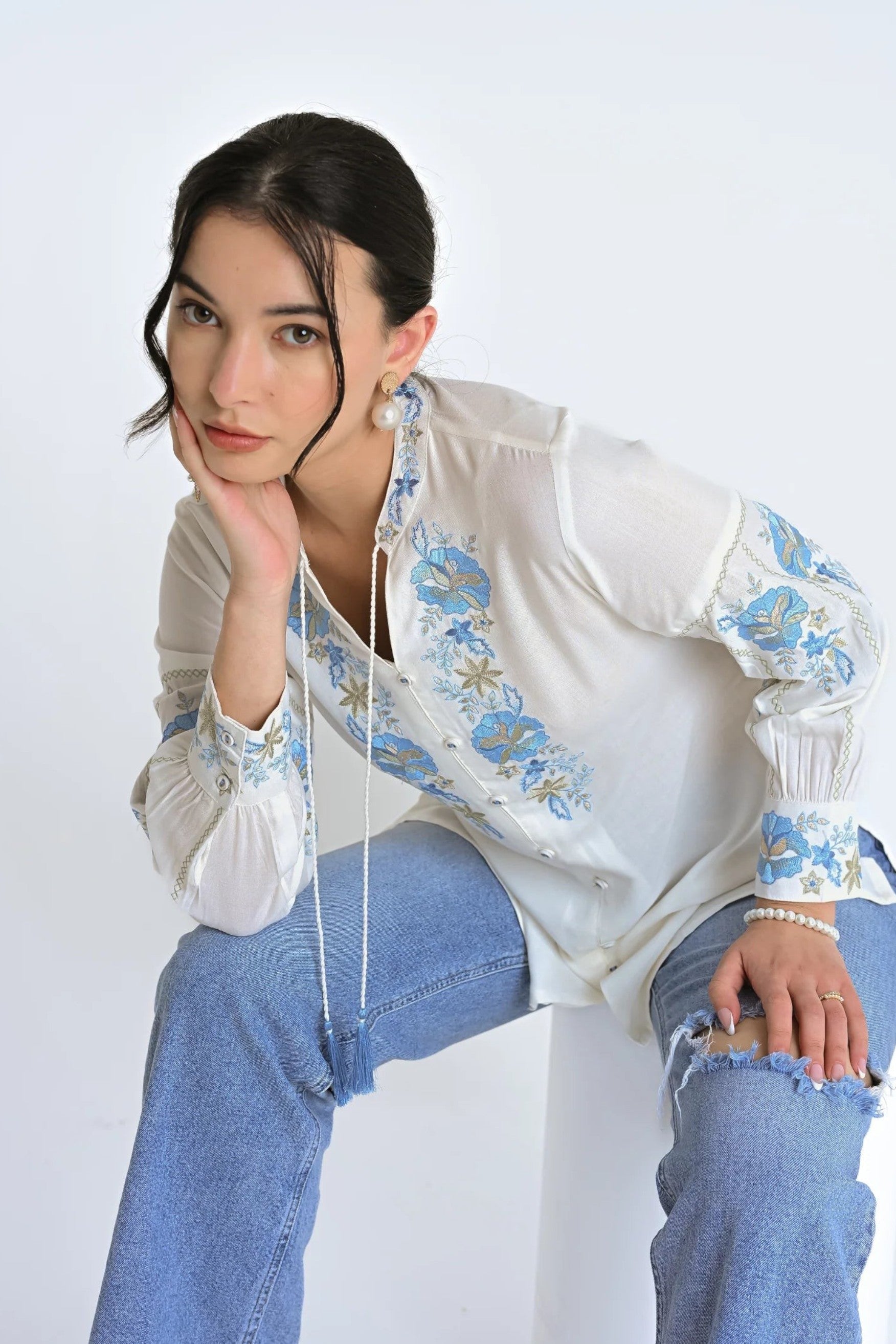 Model wearing an indigrey white shirt with blue floral embroidery, tassel details, and long sleeves.